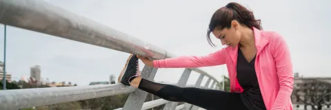 Mujer con ropa deportiva rosa y negra estirando la pierna sobre una barandilla al aire libre.